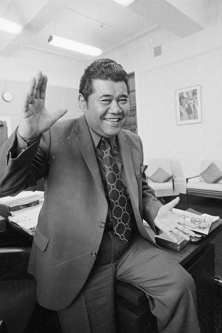 A smiling man in a suit sitting on a desk waving his hands