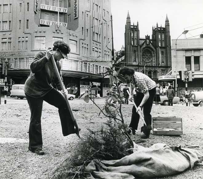 Two people in a city digging with shovels and a sack containing young trees on the ground beside them