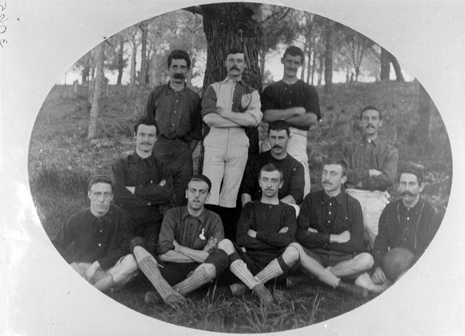 A group of ten male players standing and sitting together under a tree, many with moustaches, wearing uniforms that do not match