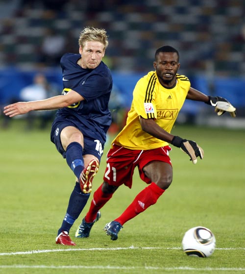 Two men during a football game, one wearing a blue uniform and kicking for a football, and one wearing a red and yellow uniform, wearing gloves.