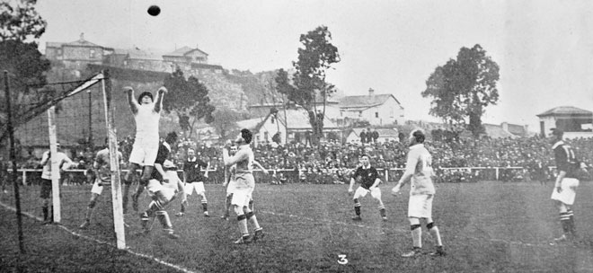 A football game watched by a crowd seated outdoors. A man jumps high in the air on front of a goal with a football above him. Other players watch him and the ball.