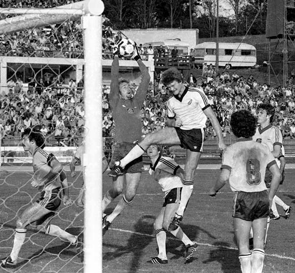 Footballers collected in front of a goal during a football game. A goalkeeper leaps to catch the ball while another jumps to head it.