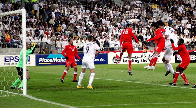 Men in white New Zealand football uniforms and men in red uniforms leap for the ball in front of a football goal. The game is taking place in a stadium.