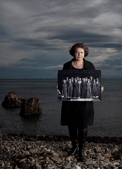 Woman standing on pebbles by sea holding an old photo of a group of women
