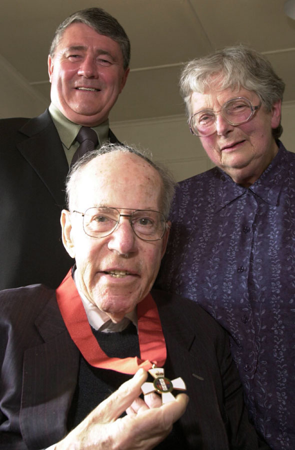Rosenberg is seated with a medal around his neck, with Jim Anderton and Ann Rosenberg behind him