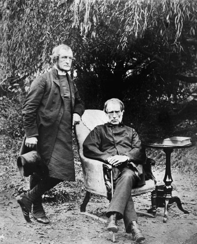 A photograph of two men under a tree, one standing and one seated
