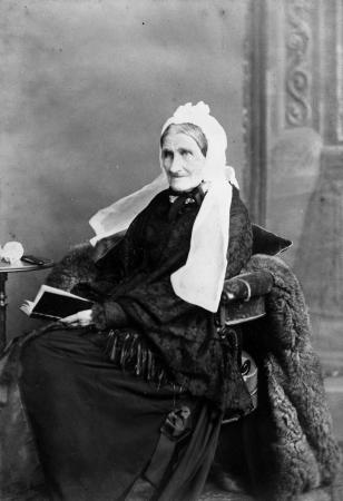 Jane Williams in later life wearing a shawl and seated in a chair holding a book.