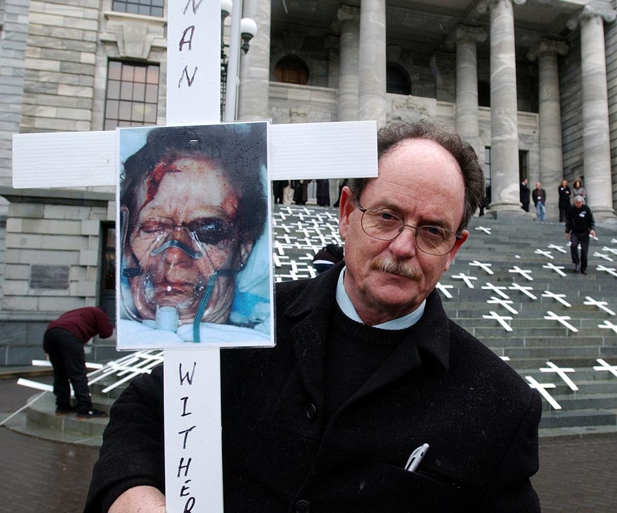 Man holding white cross with a photograph of a woman with a badly bruised face attached to it.