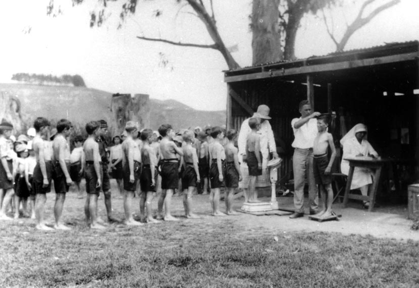 A line of children stand outside, while doctors and nurses weigh and measure others. 