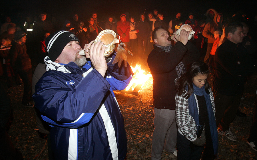 Crowd gathered around a fire at night with two men blowing conch shells in the foreground.