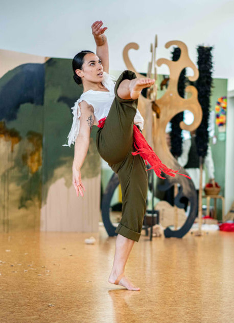 Māori woman dressed in green trousers and white top standing on one leg in an expressive pose.