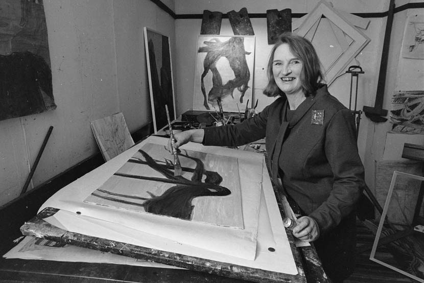Avis Higgs seated at a painting table, poised with a paintbrush over an artwork. There are other artworks, equipment, frames and mounts in the background.