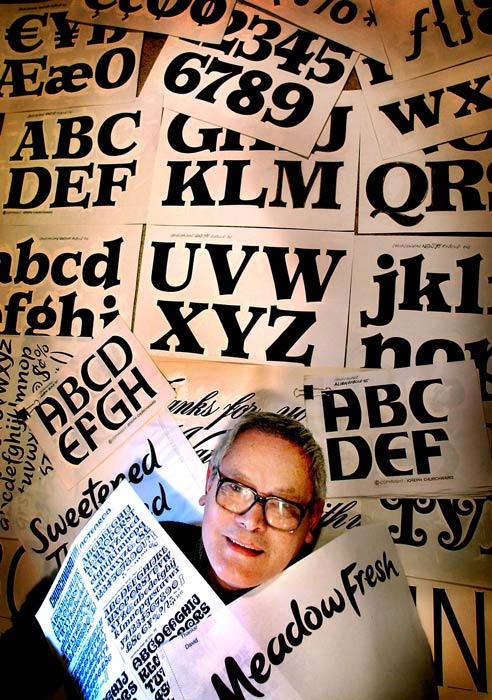 A photograph of Joseph Churchward lying on the floor surrounded by printed samples of his typefaces