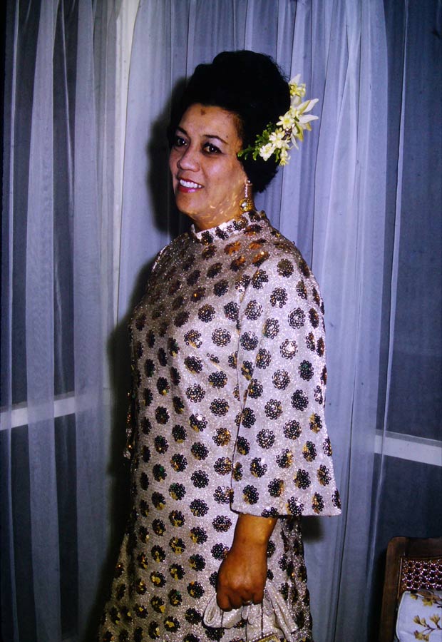 A three-quarter length photograph of Kura Ensor in a formal evening dress, with circular patterns of sparkling gold sequins. She wears flowers in her hair.