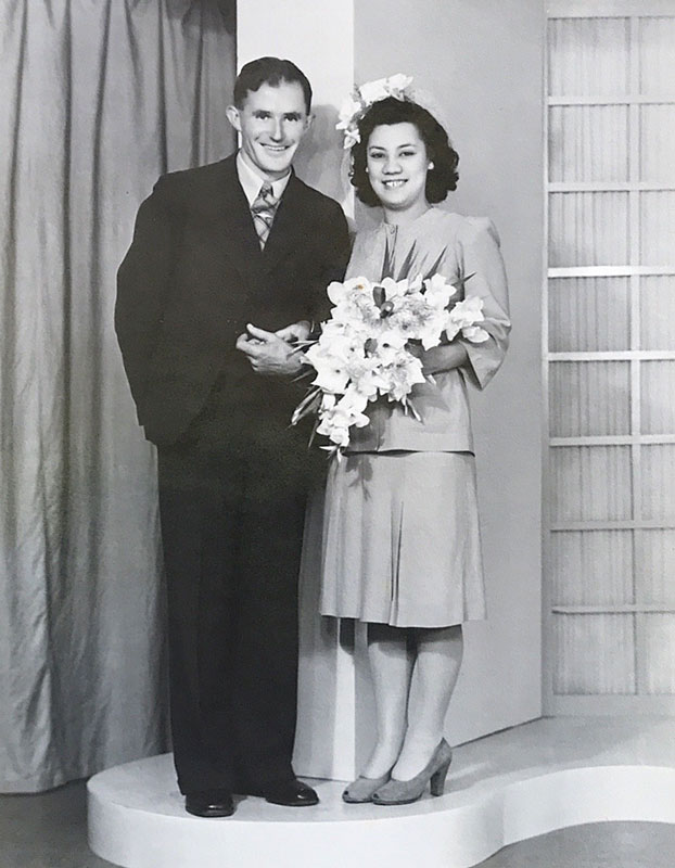 Don and Kura Ensor, dressed formally, standing in a photographer’s studio. Kura is holding a bouquet of lilies. Don is Pākehā and Kura is Māori.