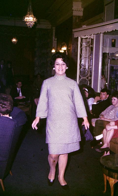 Kura Ensor on a fashion show catwalk, wearing a high-necked dress decorated with wavy black lines against a white background.