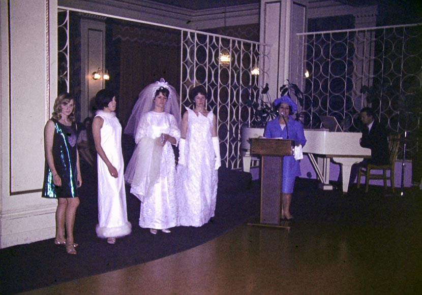 Kua Ensor in a blue jacket, skirt and hat behind a podium with models in bridal fashion standing beside her.