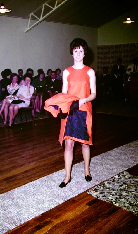 Model wearing a coral-coloured shift dress holding matching jacket with blue lining at a fashion show.