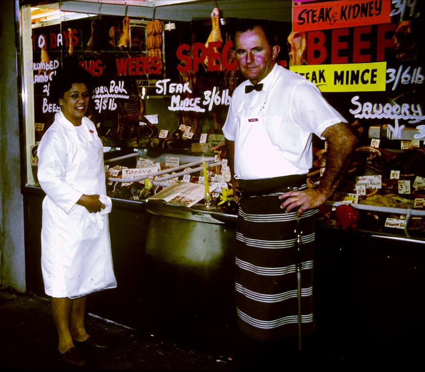 Kura and Don Ensor standing outside their shop, with meat visible through the window which has prices and advertising painted on it.