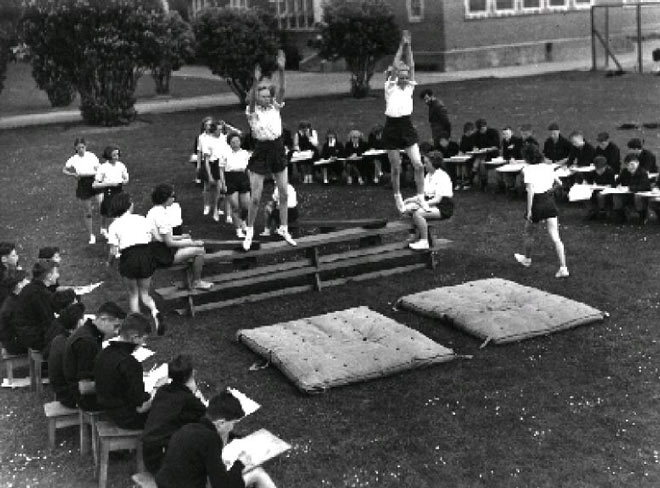 School students jump onto mattress, surrounded by a group of seated students with papers and pencils.