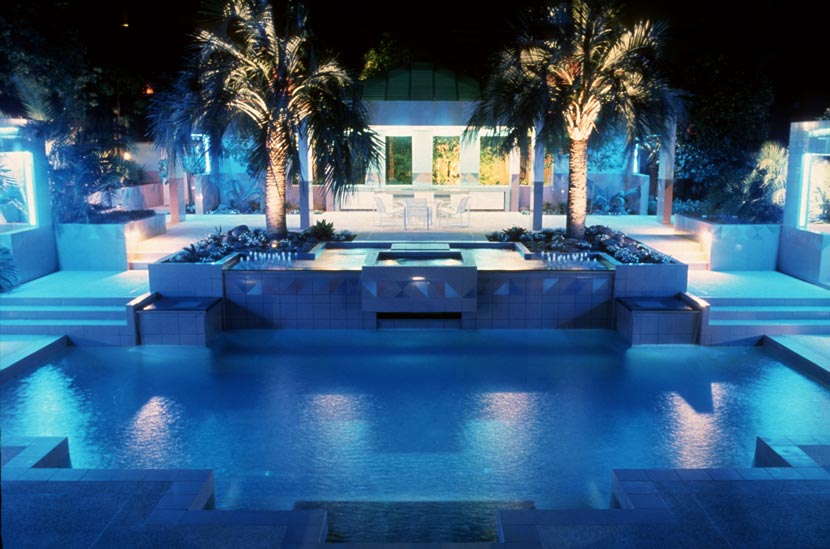 A photograph showing a garden with rectangular pool surrounded by steps and boxed gardens and lit with blue light