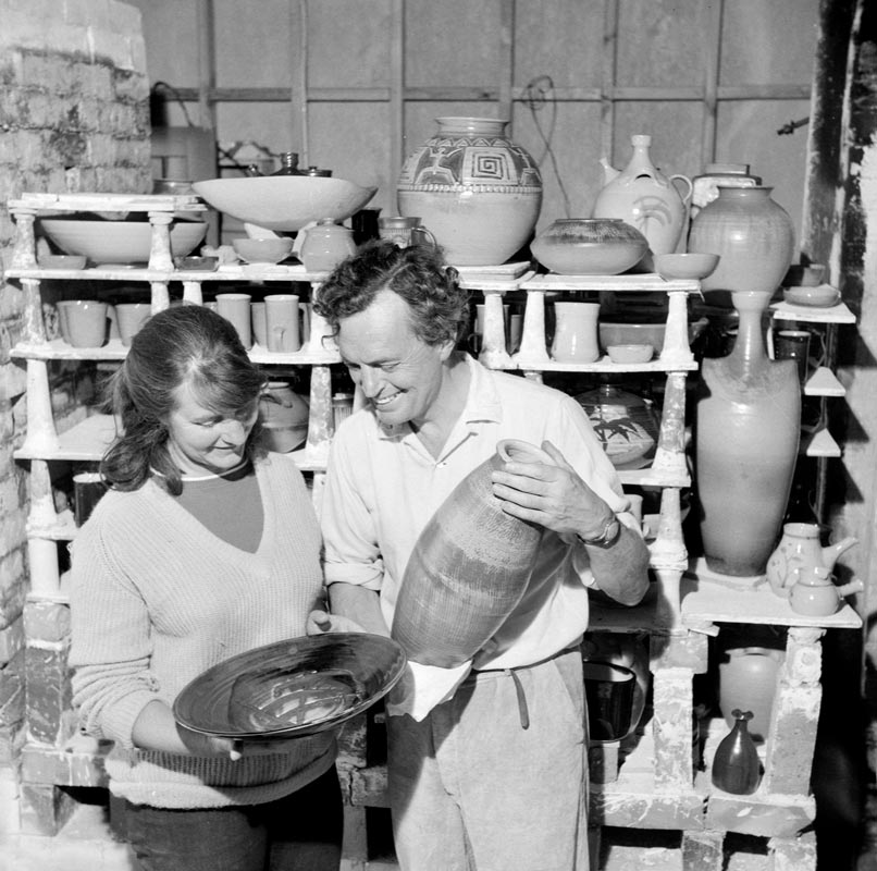 Peggy and Jack Laird in middle age, standing in the pottery workshop with a variety of completed pottery items. 