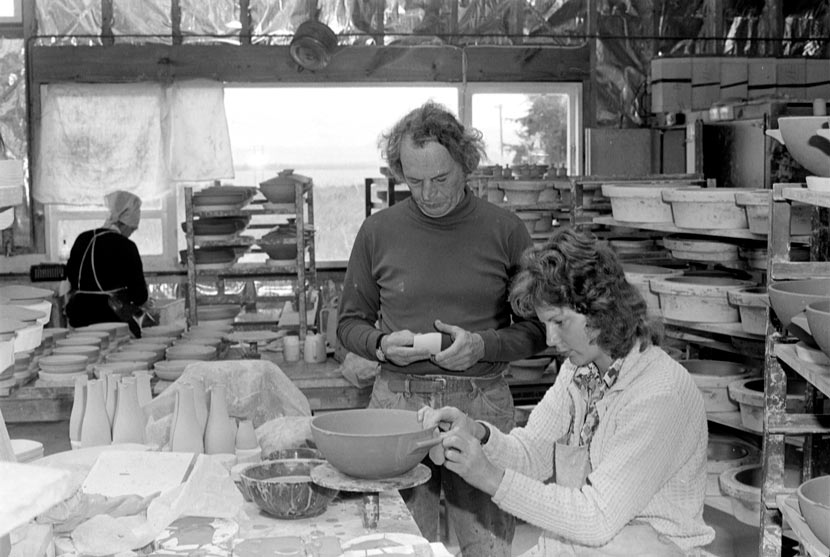 Jack Laird in middle age, standing in the pottery workshop with a variety of pottery items at various stages of completion. With him are two female workers, one of whom appears to be applying a handle to a bowl.