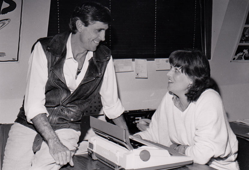 Murray Ball is sitting on a desk at which Pam sits with a typewriter in front of her. They are smiling at each other. 