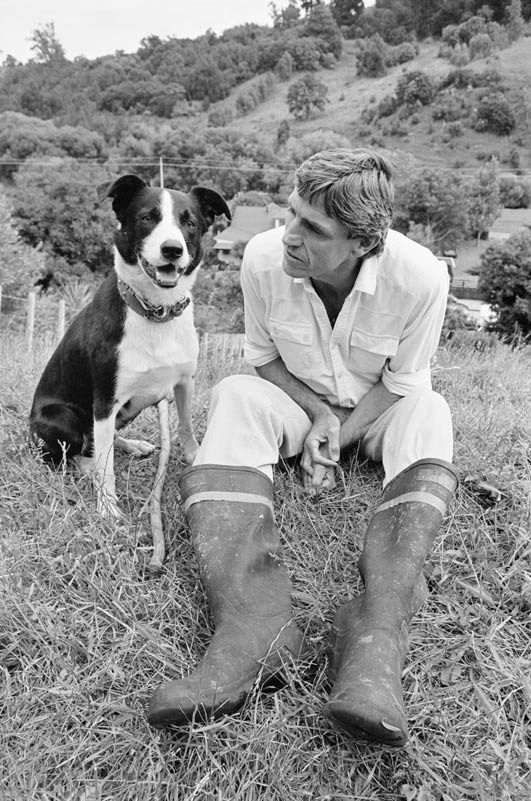 Murray Ball seated on a grassy bank beside a black and white border collie dog.
