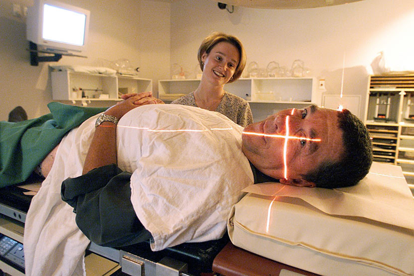 Paul Holmes lies on a hospital gurney with a blanket covering the lower half of his body, with a nurse looking on.
