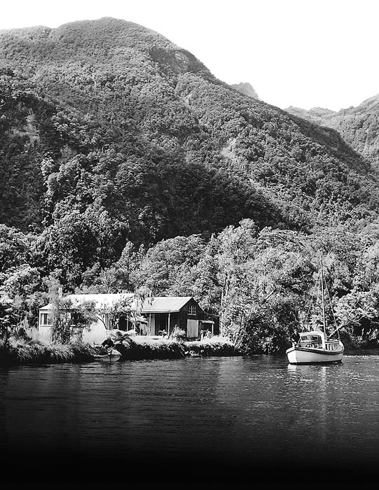 Photograph of a remote, forested, mountainous location beside a body of water. A single storey building stands beside the water, and there’s a small ship moored nearby.