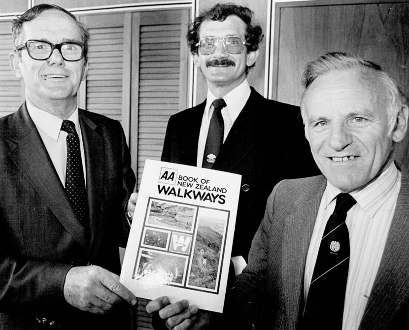 Three men in suits stand side by side, smiling at the camera, and holding a copy of the AA book of New Zealand walkways.