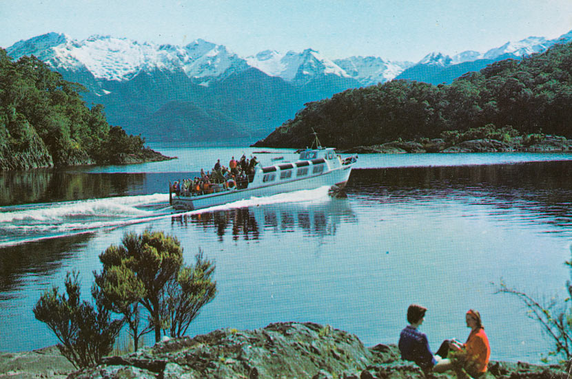 A colour image showing a body of water surrounded by forested and snow-capped mountains, with a motor vessel in the centre, heavily laden with passengers. Two people picnic in the right foreground. 