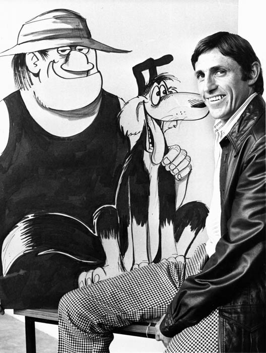 Murray Ball as a young man, seated beside an enlarged illustration of the Wal’ and Dog ‘Footrot Flats’ characters. 