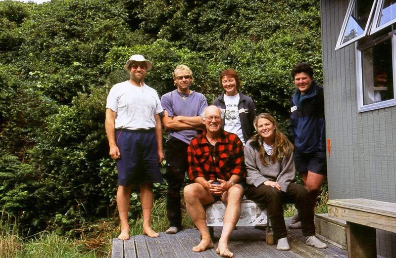 Six people pose for the camera on the deck of a hut, four standing and two seated.