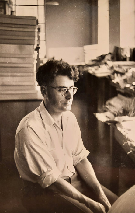 Tony Druce as a young man, seated indoors and surrounded by boxes and files. 