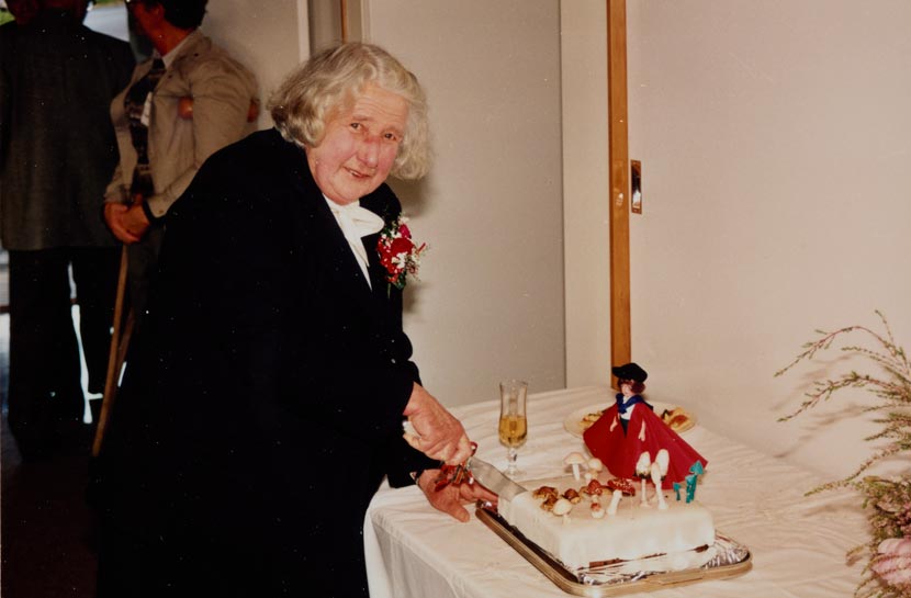 An elderly woman cutting a cake topped with icing mushrooms.
