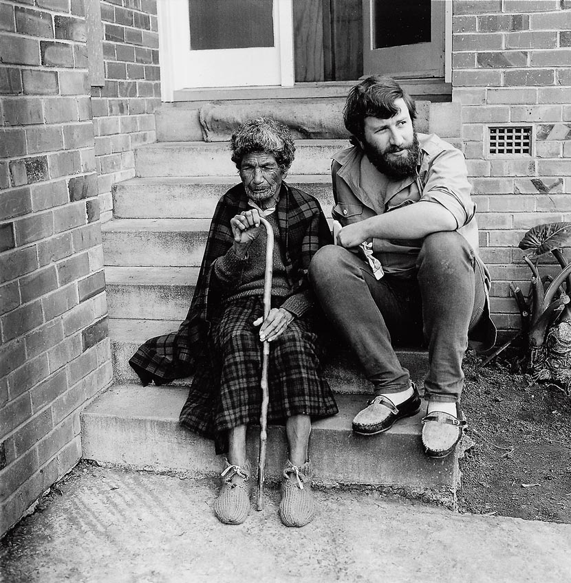 Michael King as a bearded young man, seated on concrete steps outside a house with Ngakahikatea Whirihana, an elderly Māori woman with a moko (facial tattoo). 