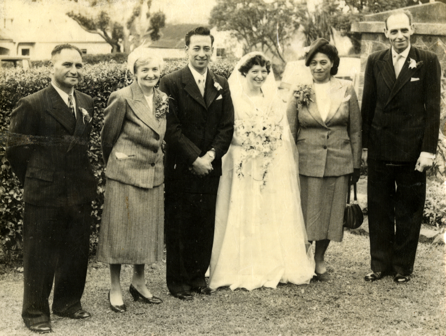 A posed wedding photograph in an outdoors setting, showing the bride and groom flanked by their respective parents. All are formally dressed. Deirdre, who wears a wedding dress, holds a bouquet of flowers. 