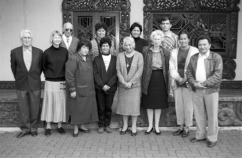 A group of 12 people standing in front of a building with the doorway and window lintels carved in Māori designs. 