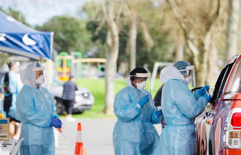 Colour photograph of three people in medical personal protective equipment standing next to a car.