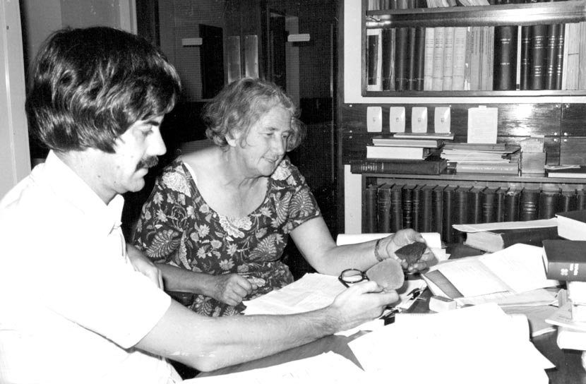 A man and woman seated at a table covered in papers and books.