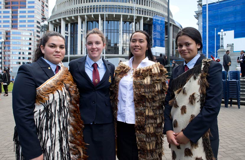 Four girls in school uniform, three wearing kākahu woven cloaks, in front of the Beehive in Wellington