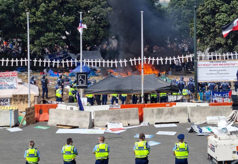 Police in high-visible clothing and riot gear stand in front of grass area littered with rubbish.