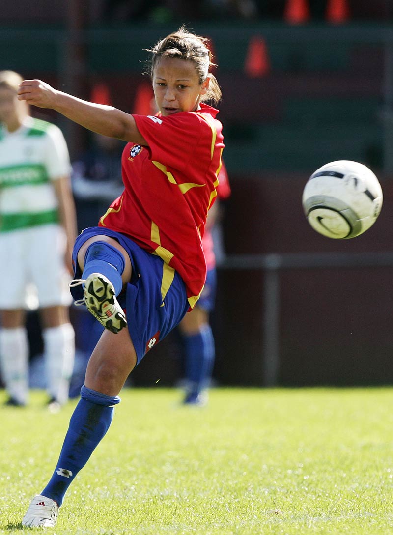 A young woman in a red and blue football uniform kicks a ball towards the front of the photograph.  