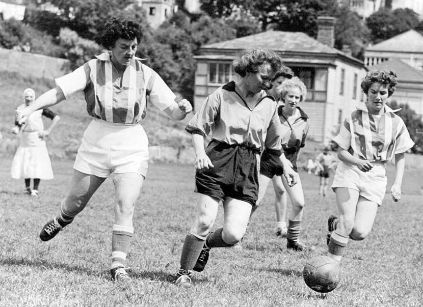 A woman in a football uniform running during a football game, with a football in front of her. She is chased by two players in a different uniform.