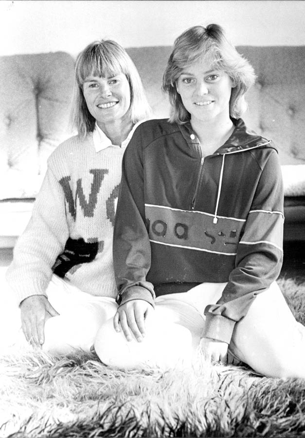 A young adult women sits with an older woman behind her, both in casual clothes, in front of a couch.