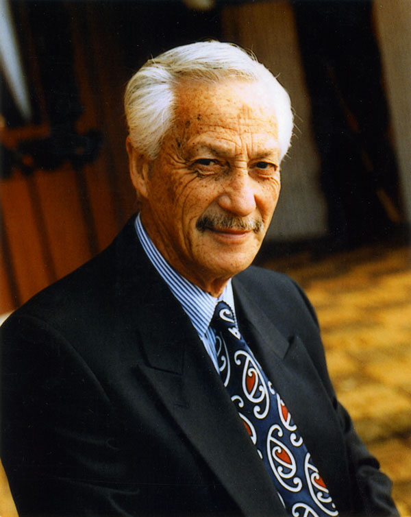 A head and shoulders portrait taken in later life, with Hugh Kawharu in a suit jacket and a tie decorated with Māori designs. He’s seated in front of Tumutumuwhenua, the wharenui (meeting house), at Ōrākei.