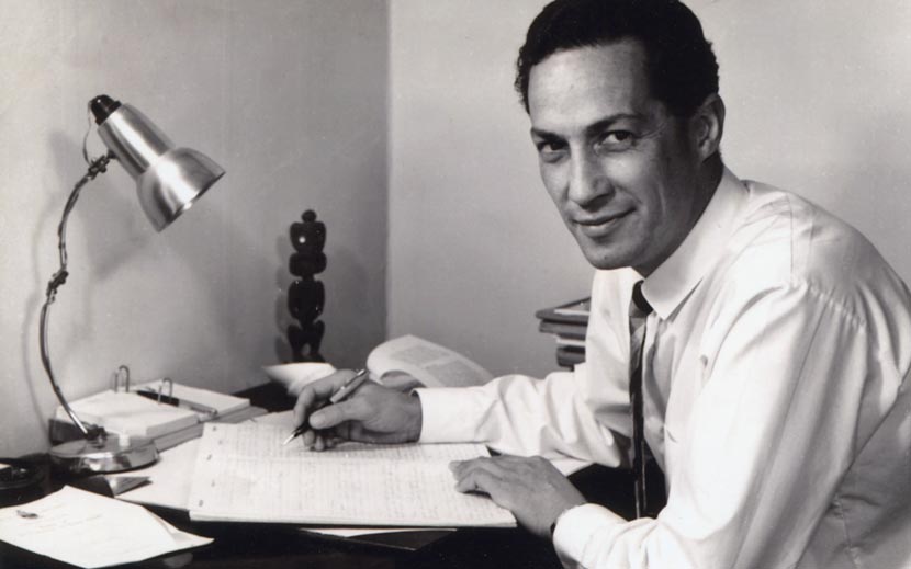 Hugh Kawharu as a young man, seated at a desk with a lamp, papers, and a small carved Māori figurine. 