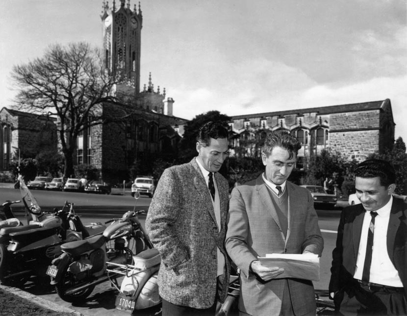 Three men standing side by side and looking at some papers, with the University of Auckland buildings behind them.